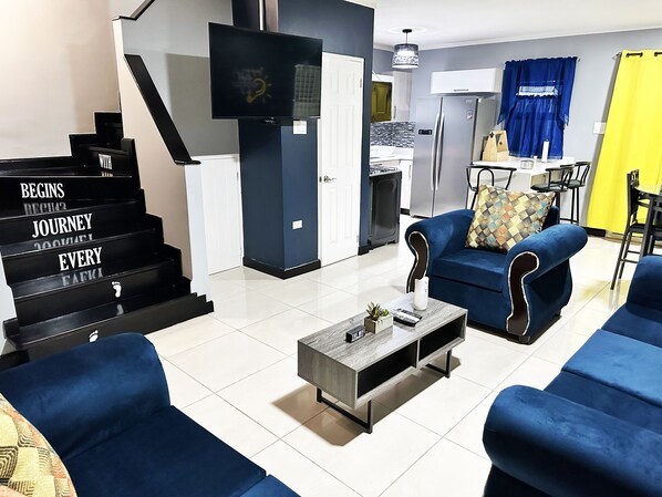 A modern living room with a staircase, a flat-screen TV, a dining area, and a kitchen.
