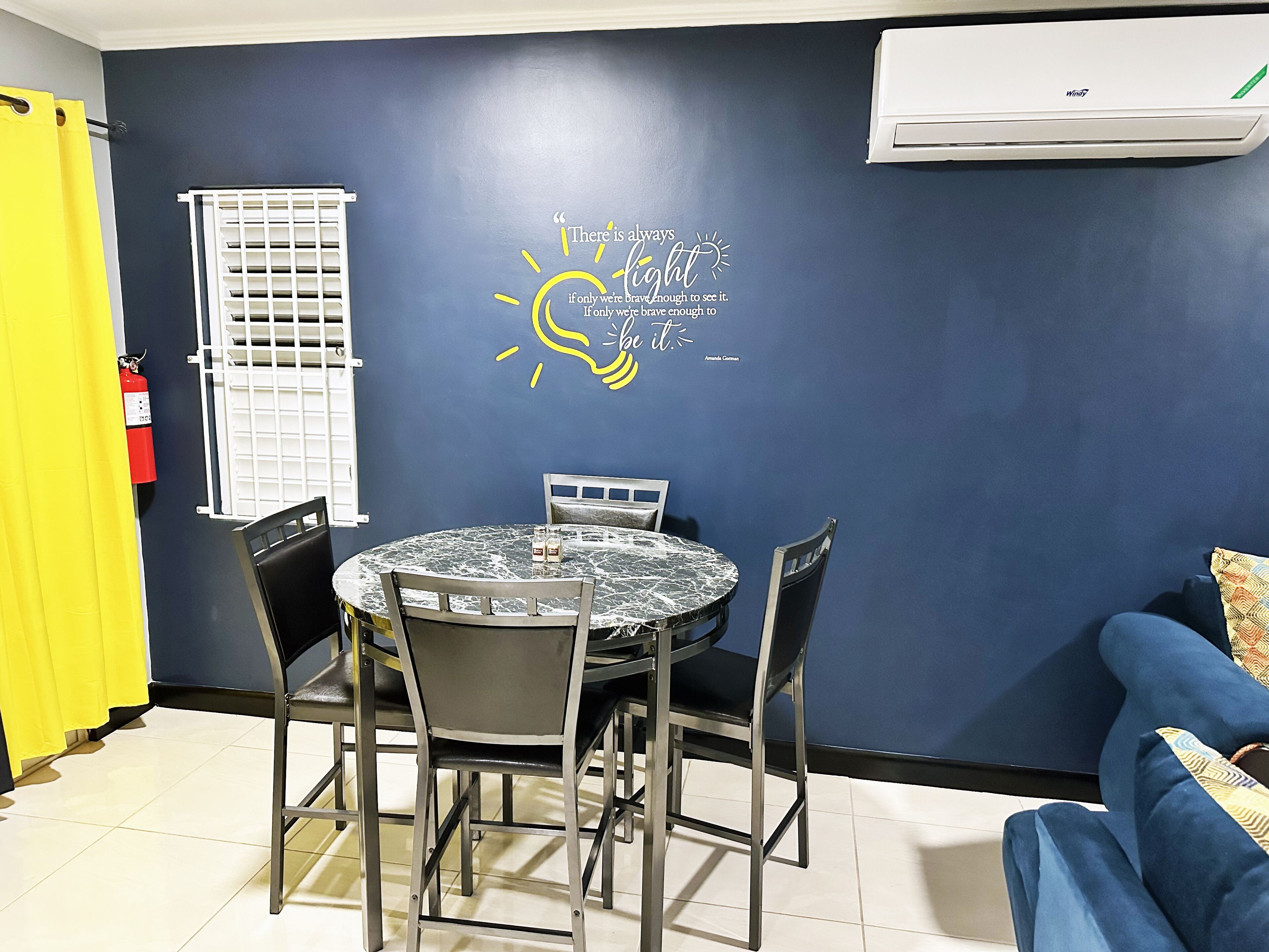 A room with a round table, two chairs, a blue wall with a motivational quote, a white air conditioning unit, and a yellow curtain.