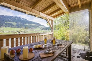 Een houten tafel gedekt met eten en drinken, een houten bank, en uitzicht op een bergachtig landschap.