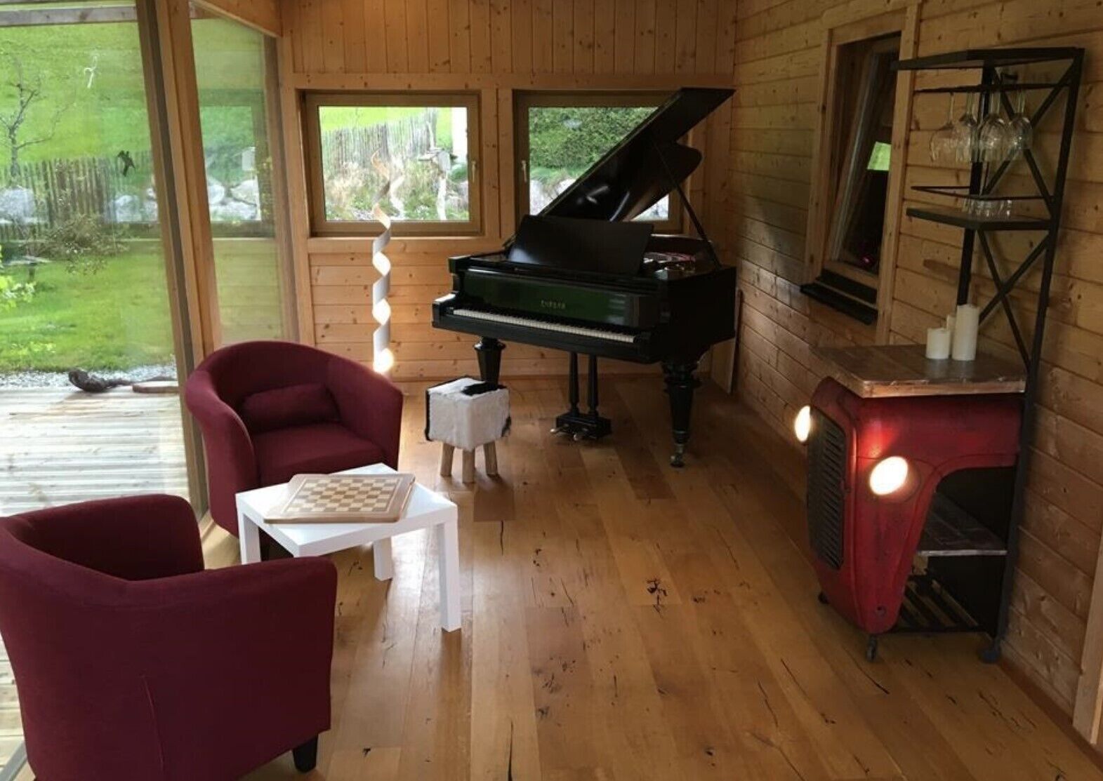 A wooden cabin interior with a grand piano, red chairs, a small table with a chessboard, and a glass display case.