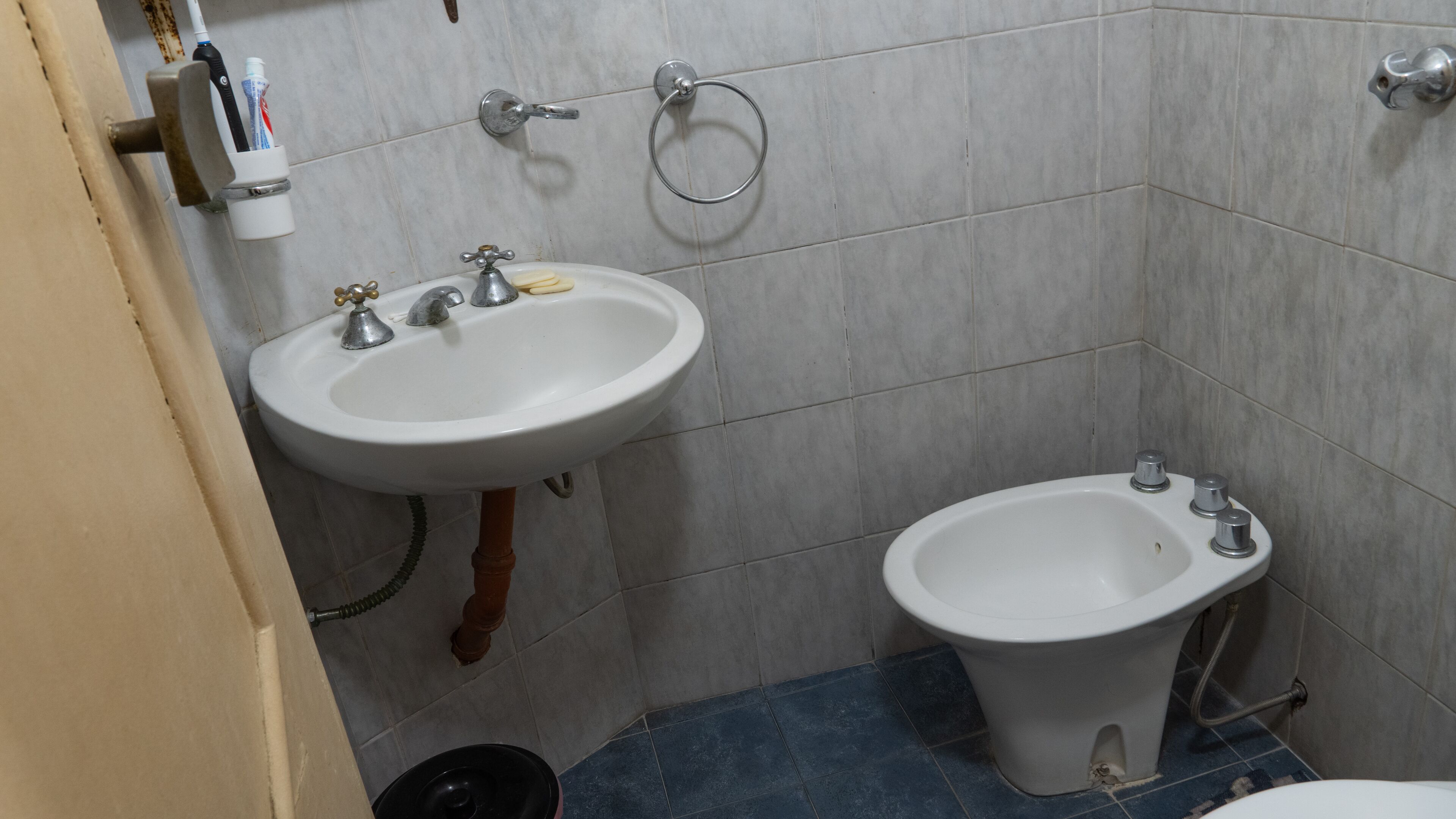 A bathroom with a white sink, a bidet, and a toilet. There are two faucets on the sink and a towel ring on the wall.