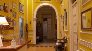 A hallway with yellow walls, white trim, and a checkered floor. There are framed pictures on the walls, a clock, and a lamp on a table. - Sevilla home Hotel (Buenos Aires)