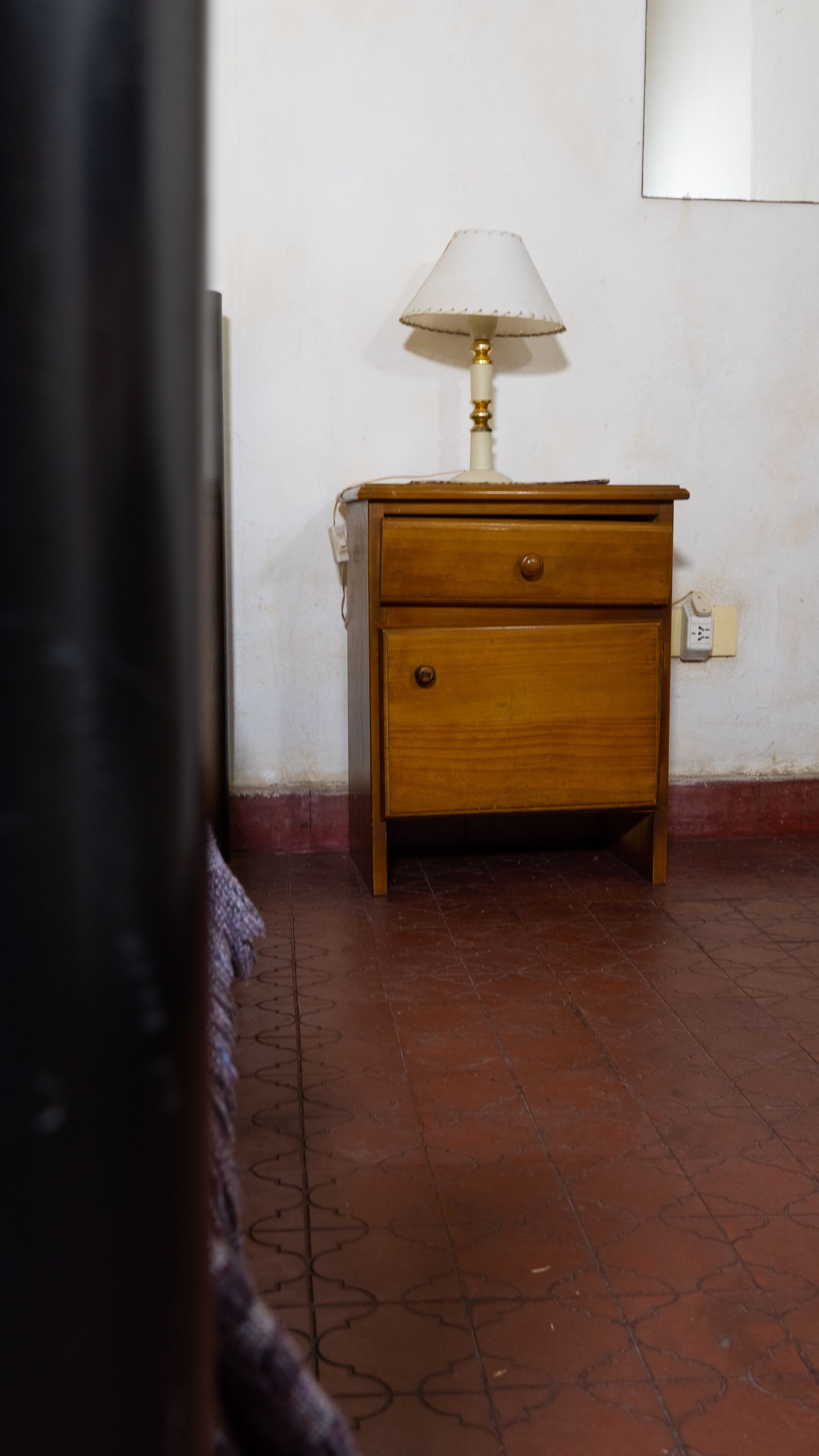 A wooden nightstand with a lamp, a drawer, and a power outlet on a tiled floor.