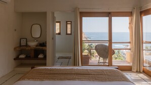 Quarto com cama, mesa de cabeceira de madeira, grande janela com vista para o oceano e varanda com cadeira e planta em vaso.