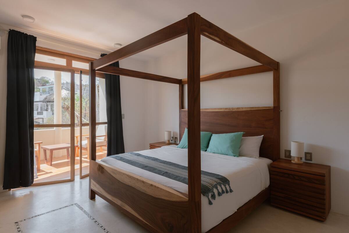 Una cama con dosel de madera, con cubrecama blanco y almohadas verde azulado. Junto a la cama, una mesita de noche de madera con una lámpara.