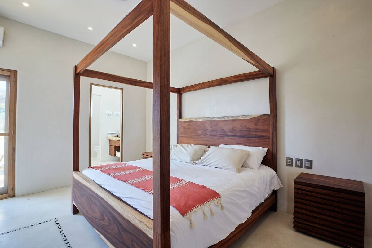 Una cama con dosel de madera con ropa blanca, un mesita de noche de madera y un espejo en la pared.
