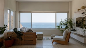 Uma sala de estar moderna com uma grande janela com vista para o mar, móveis de madeira e plantas em vasos.