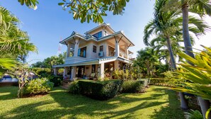 A large, two-story house with a covered porch, surrounded by lush greenery and palm trees. - Pool Villa with maid service- walk to white sand beach! (Paknampran)