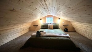 Un dormitorio acogedor y pequeño en el ático, con techo inclinado, una cama individual, un ventanal y dos lámparas fijadas a la pared.