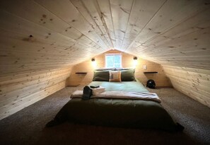 Une petite chambre mansardĂ©e, chaleureuse et accueillante, avec un plafond en pente, un lit simple, une fenĂȘtre et deux lampes fixĂ©es au mur.