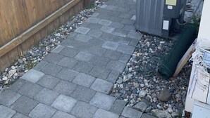 Un patio trasero con cerca de madera, un patio empedrado y una unidad de aire acondicionado grande.