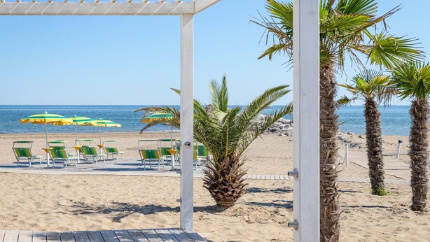 A beach view with a wooden pergola, palm trees, beach chairs, and umbrellas. - Nice home in Cavallino-Treporti (Cavallino-Treporti)