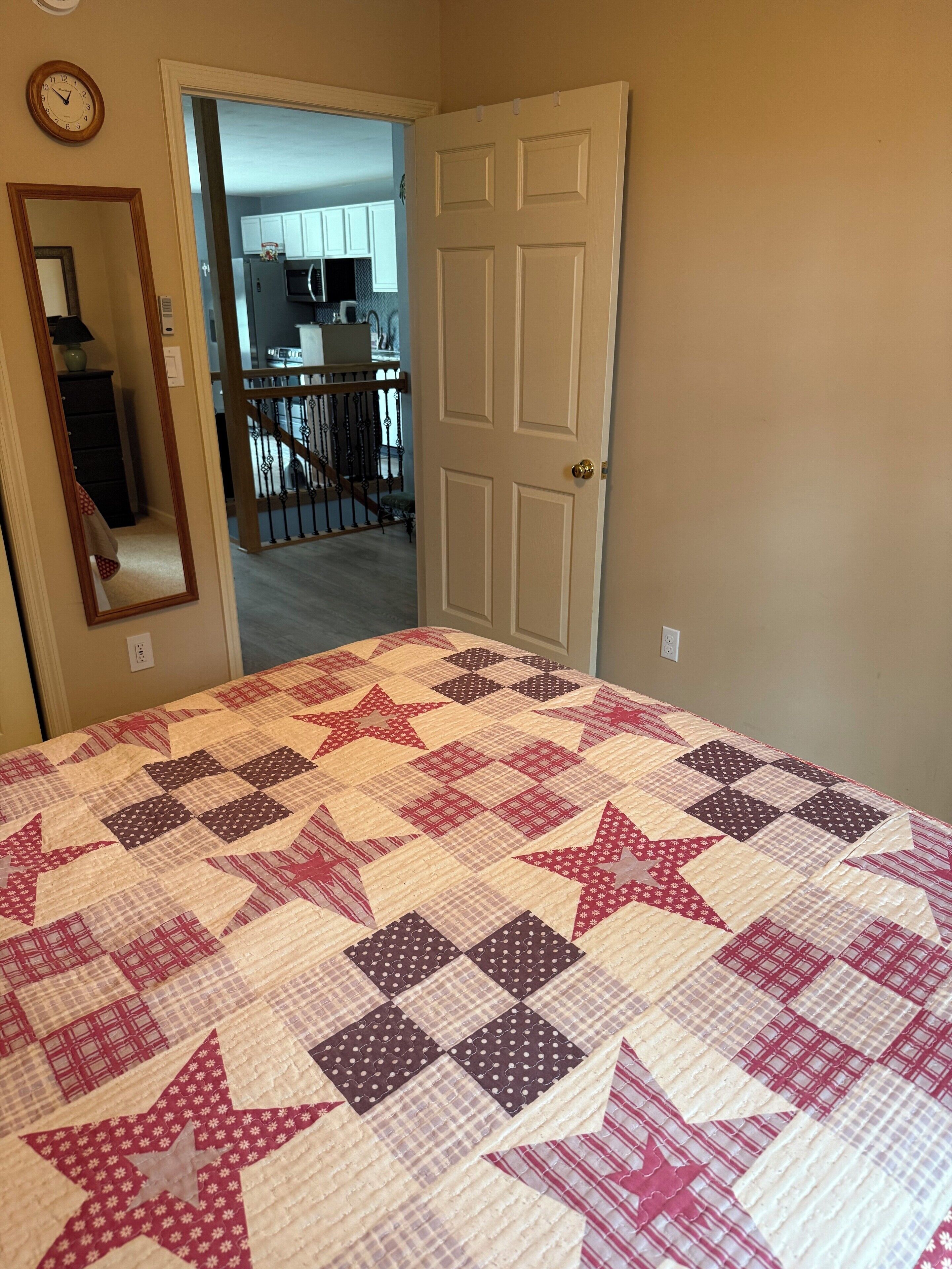 Ein Schlafzimmer mit Bett, einer Tür, einem Spiegel und Blick in einen Küchenbereich.
