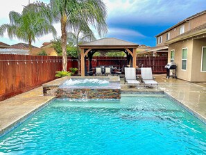 Une piscine avec un jacuzzi, entourée d’une clôture en bois, un patio couvert et une maison en arrière-plan.