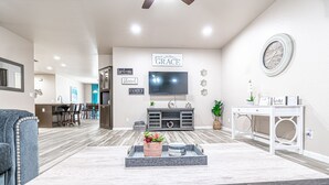 Una sala de estar moderna con un televisor de pantalla plana, un sofá gris, una mesa de centro con objetos decorativos y un comedor al fondo.