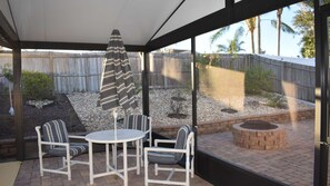 Een overdekt terras met een tafel en stoelen, een gestreept parasol en uitzicht op een achtertuin met een houten schutting en beplanting.