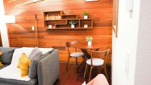 A room with a wooden wall, a grey sofa with a yellow pillow, a small table with chairs, and shelves with plants and decorative items.