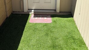 Uma porta branca com um tapete de entrada em frente, localizada num canto com um pequeno trecho de grama sintética.