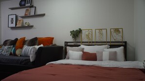 Quarto com cama, sofá e prateleira na parede com quadros e planta.
