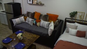 Uma sala de estar com sofá, mesa de centro e cama. Há também uma geladeira e uma estante com objetos decorativos.