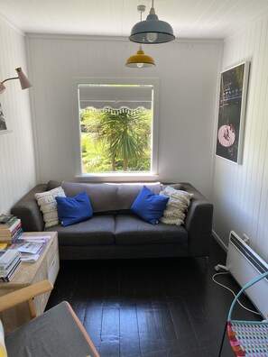 A cozy living room with a grey sofa, blue cushions, a wooden coffee table with books, a window with a view of greenery, and a pendant light. - Rosie's cottage in Stanmore Bay , a short walk to beach with a great seaview. (Whangaparāoa)