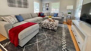 A modern living room with a grey sectional sofa, a patterned ottoman, a flat-screen TV, and a ceiling fan.