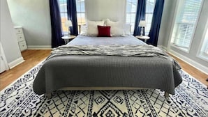 A bedroom with a large bed, two bedside tables, a nightstand, a patterned rug, and a window with curtains.