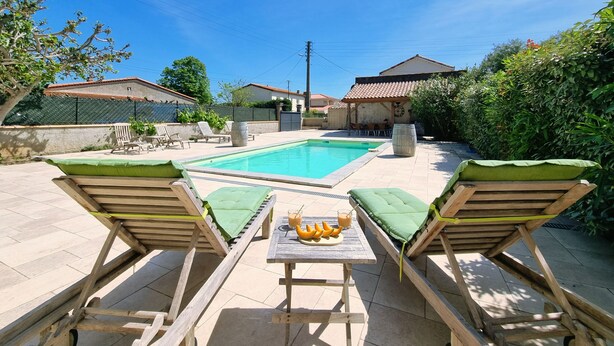 Un patio trasero con piscina, dos sillones de madera con cojines verdes, una mesa con frutero y bebidas, y un área de estar cubierta.