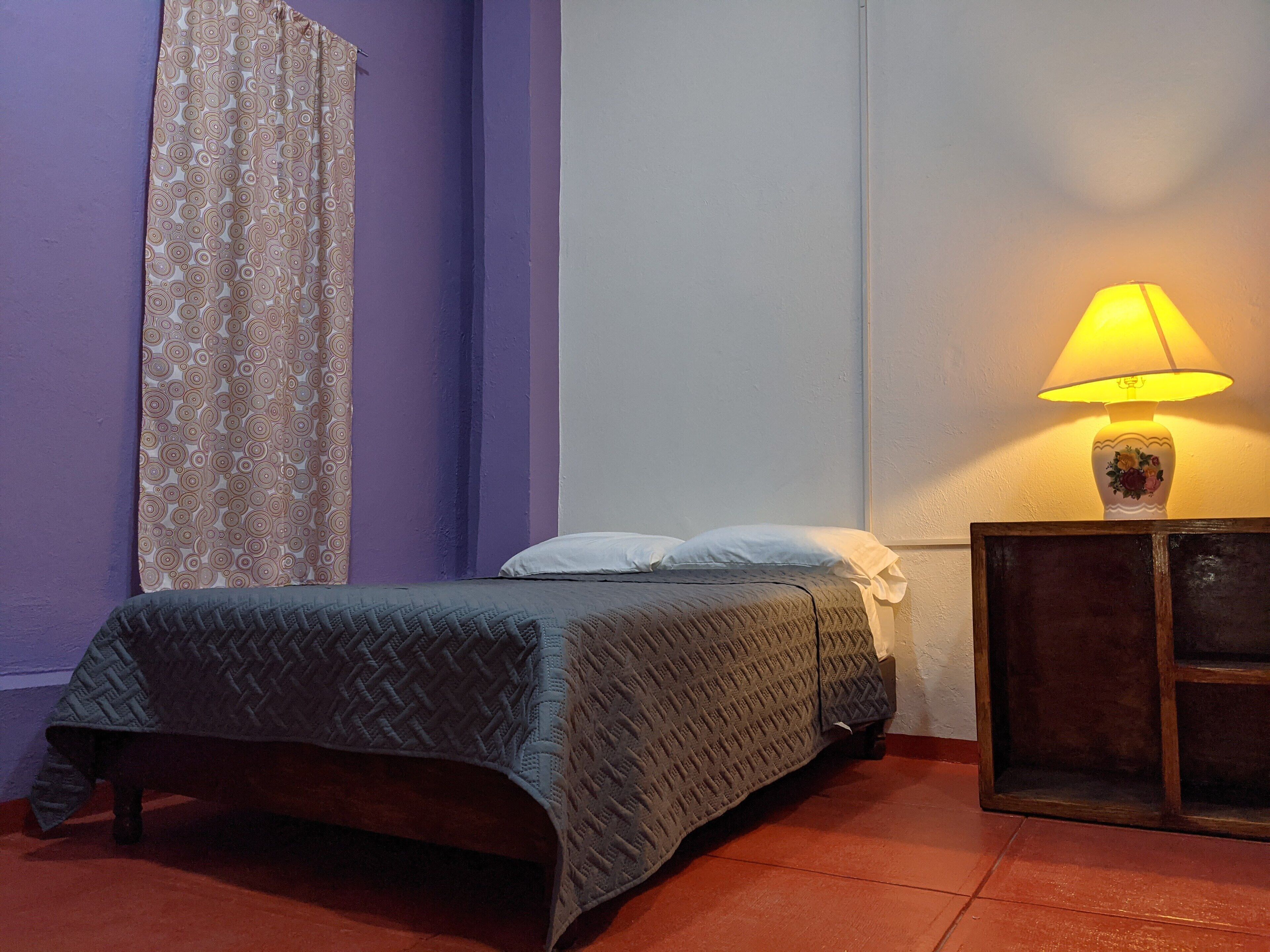 A single bed with a patterned bedspread, a bedside lamp, and a curtain on a purple wall.