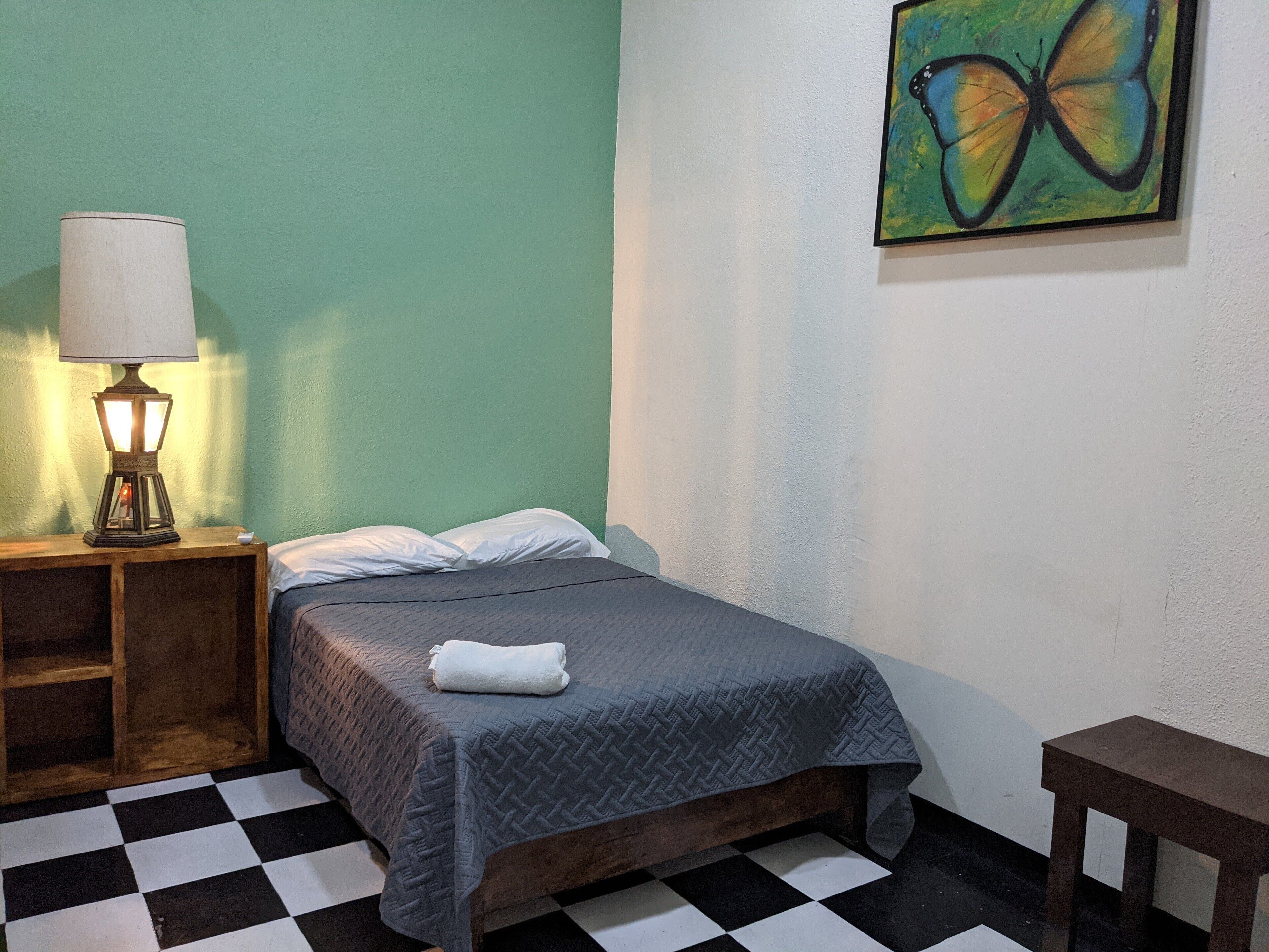 A room with a bed, a nightstand with a lamp, a painting, and a checkered floor.