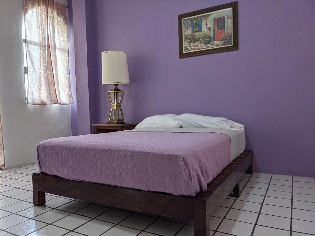 Une chambre avec un lit, une table de chevet avec une lampe, une fenêtre avec des rideaux et un tableau au mur.