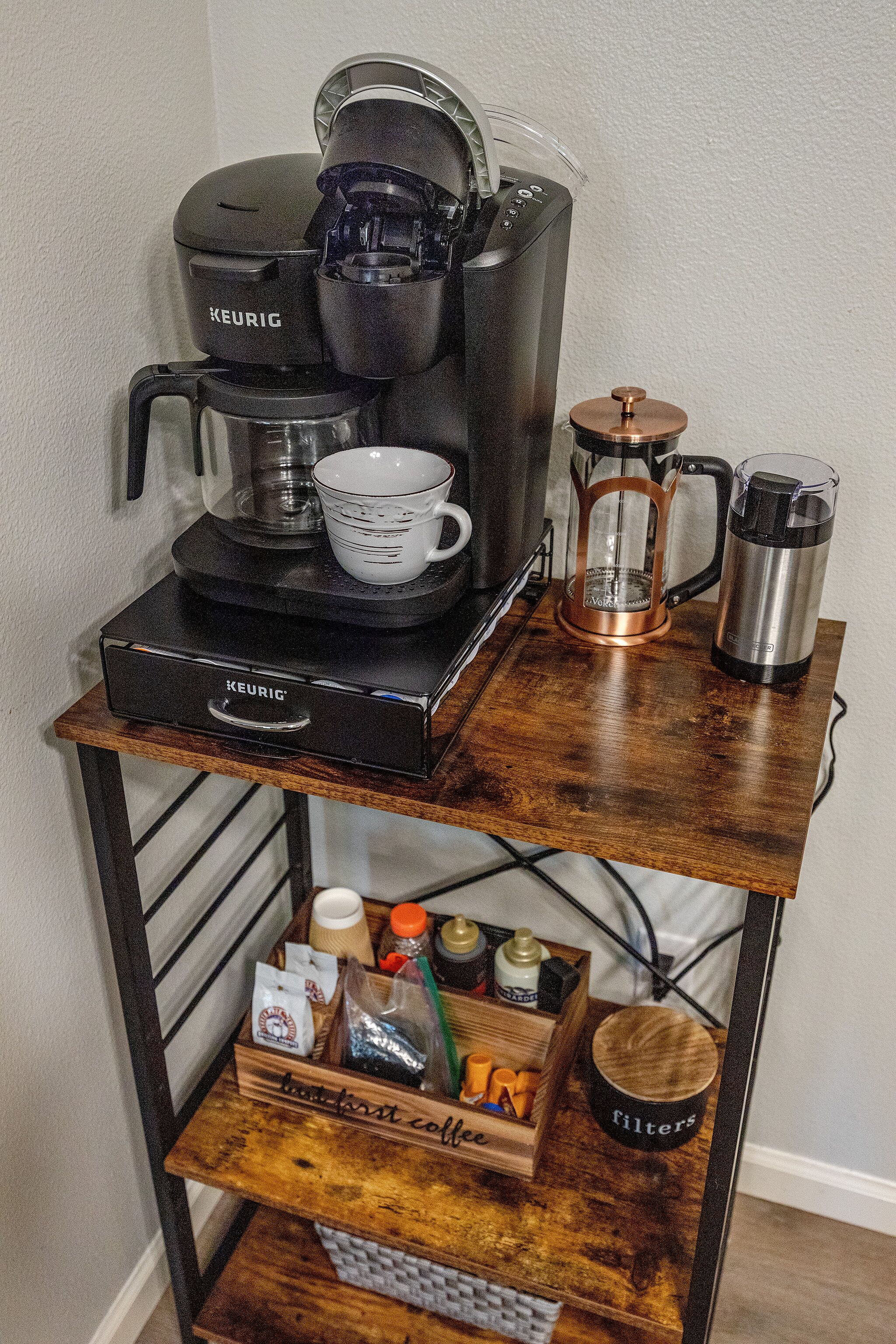 A coffee station with a Keurig machine, coffee cups, a coffee press, and a coffee grinder on a wooden table.
