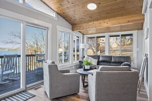 Relax in a spacious room with a high ceiling, featuring a cozy seating area and large windows offering views of the outdoors and a wooden deck.