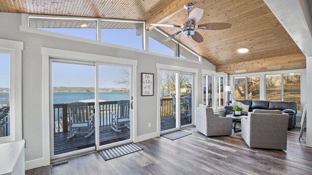 A spacious living area with a large window overlooking a body of water, a ceiling fan, and a wooden deck. - Honeysuckle Beach House - Next to Shady Gators - Private Beach! (Lake Ozark)