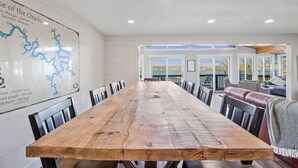 A spacious dining area with a long wooden table and chairs, a map on the wall, and a view of the outdoors. - Honeysuckle Beach House - Next to Shady Gators - Private Beach! (Lake Ozark)