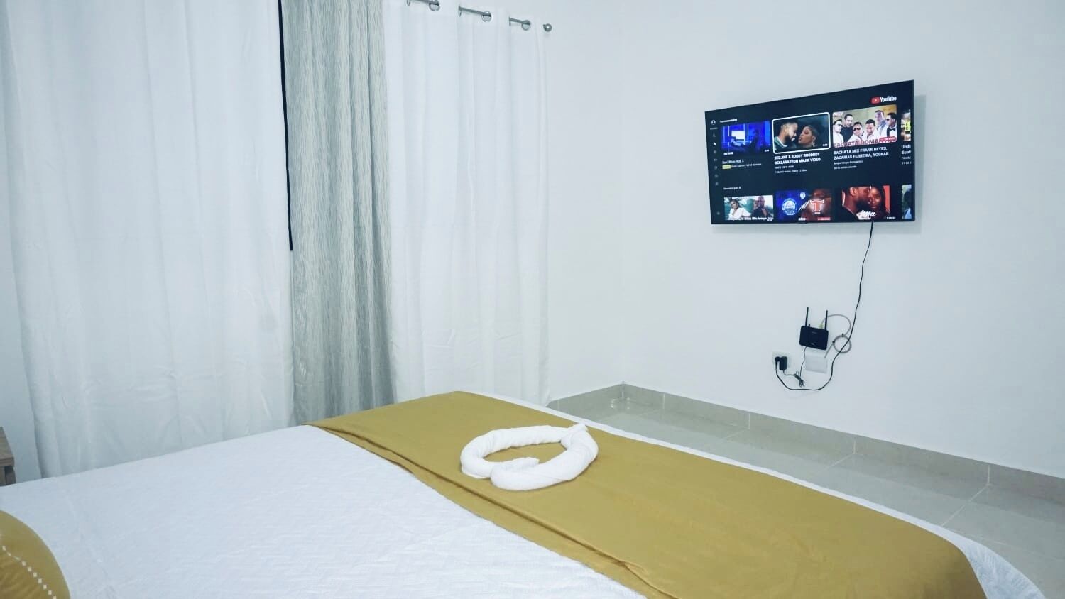 A hotel room with a bed, a wall-mounted TV displaying various channels, and a power outlet on the wall.