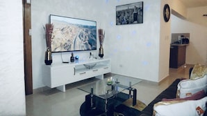 A modern living room with a flat-screen TV, white entertainment unit, glass coffee table, and a sofa with cushions. - Confidence Luxurious Apartment (Santiago de los Caballeros)