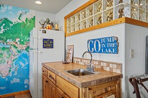 A kitchen with a map, a refrigerator, a sink, wooden cabinets, and a wall sign that reads 'GO JUMP IN THE LAKE'. - Relaxing Lakeside Getaway on Beautiful Hebgen Lake! HOT TUB with lake views- Beachside Lodge (West Yellowstone)