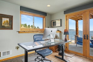 Un bureau à domicile équipé d’un bureau, d’une chaise et d’un ordinateur. Il y a une fenêtre donnant sur des arbres et une porte qui mène à un balcon avec vue sur l’eau.
