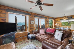 Ein Interieur einer Blockhütte mit Ledersofa, Fernseher und Essbereich mit Bergblick.
