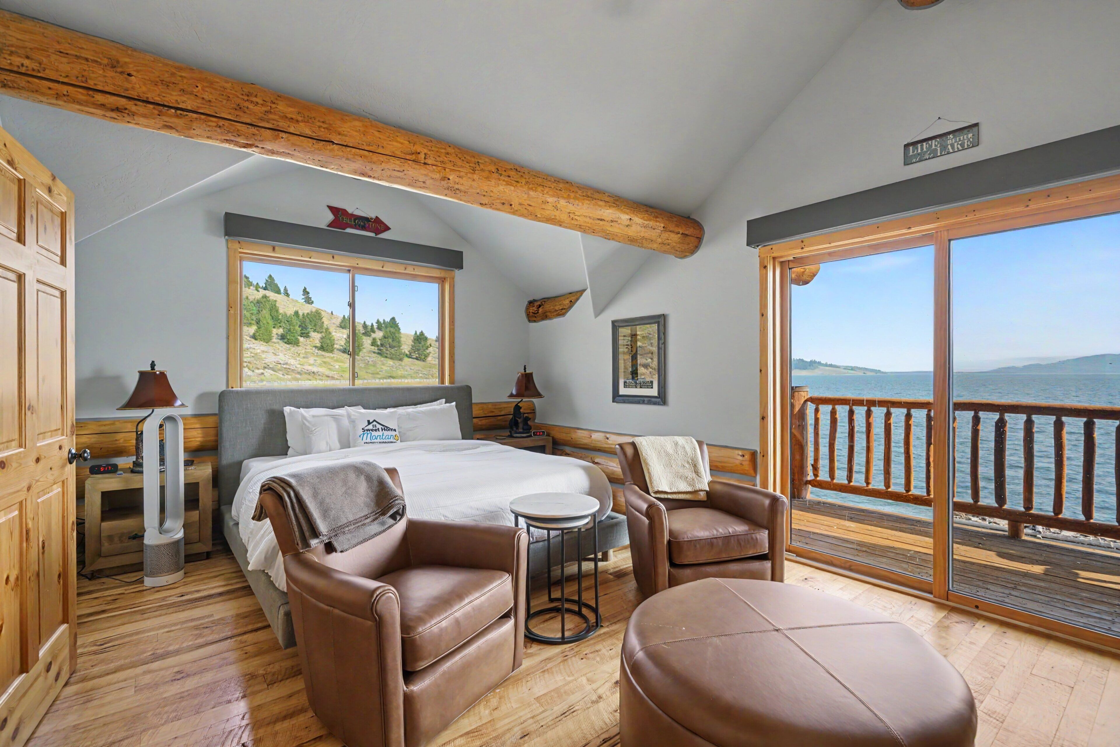 A bedroom with a bed, two chairs, a small table, a window with a view of the lake, and a wooden ceiling.