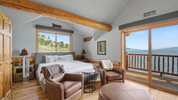 A bedroom with a bed, two chairs, a small table, a window with a view of the lake, and a wooden ceiling.