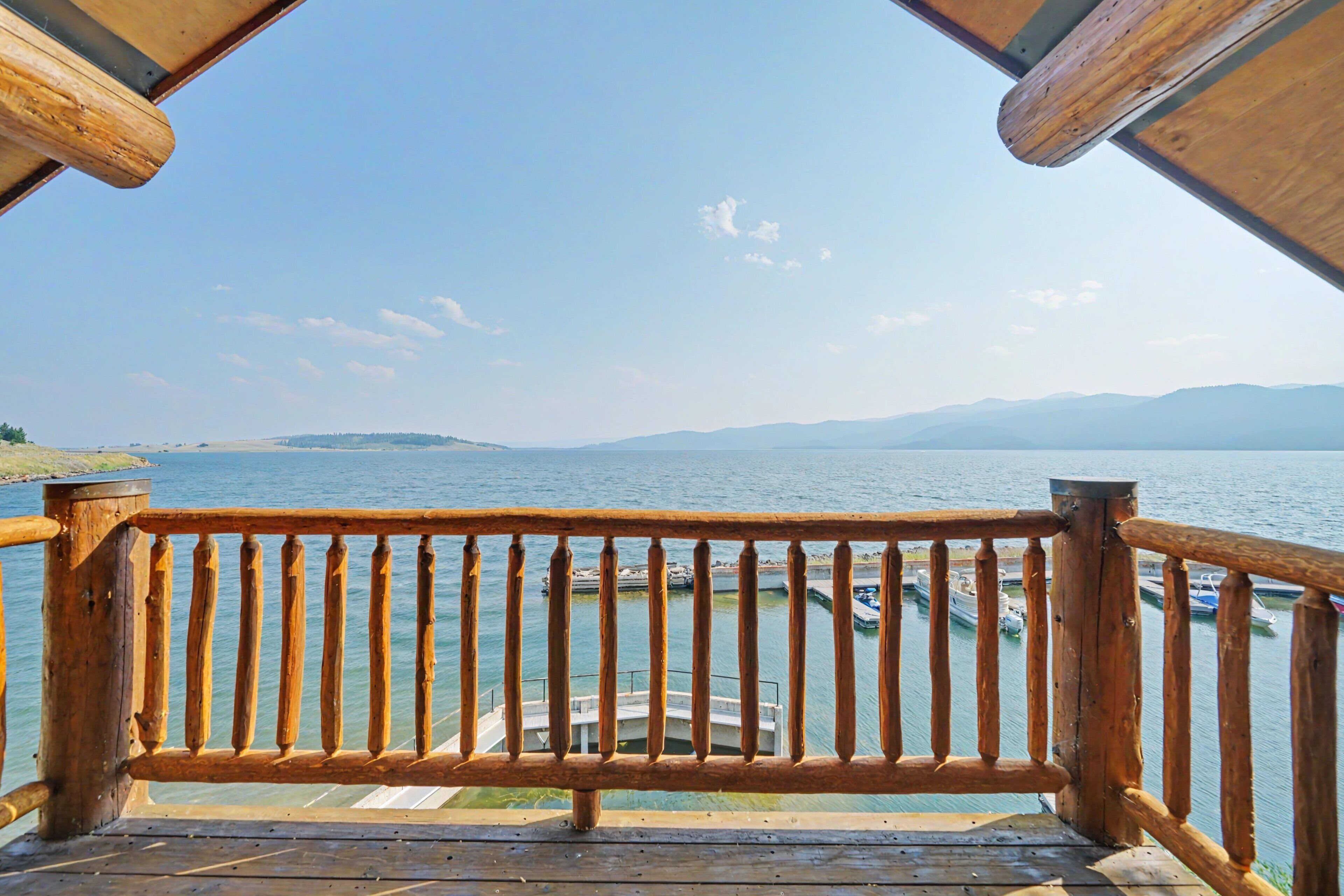 A wooden deck with a railing overlooking a calm body of water, boats docked, and a clear sky.
