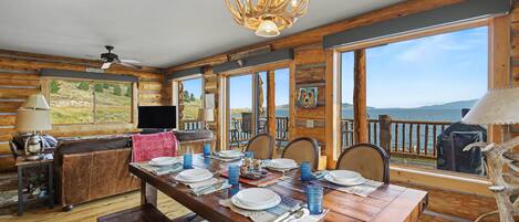Interior de una cabaña de madera con una mesa de comedor para cuatro, un televisor y una vista panorámica a través de amplias ventanas.