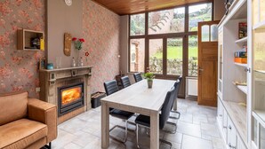 A cozy room with a fireplace, a dining table with chairs, a sofa, and a large window offering a view of the garden. - Beautiful holiday home in Dinant with garden (Dinant)