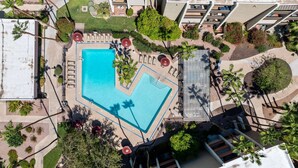 Vista aérea de um hotel com uma piscina retangular grande, rodeada por espreguiçadeiras e guarda-sóis, além de um jardim e edifícios altos ao fundo.
