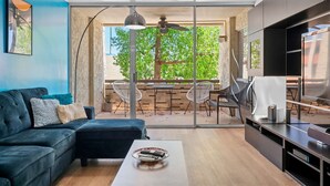 Uma sala de estar moderna com um sofá azul escuro, uma mesa de centro e uma televisão. Há um varanda com uma mesa e cadeiras, e um ventilador de teto.
