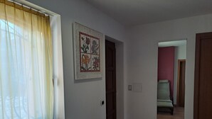 Un pasillo con una ventana cubierta por cortinas de gasa, una obra de arte floral colgada en la pared, una puerta que da a otra habitación y un piso de baldosas.