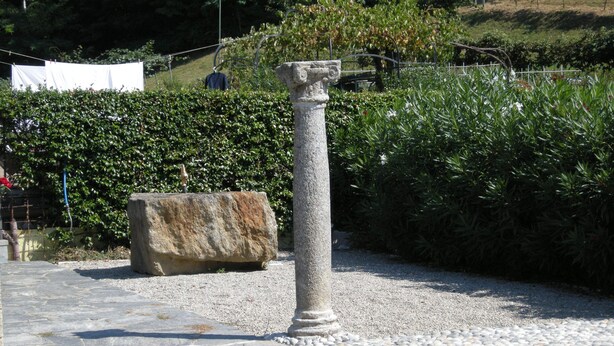 Un pilar de piedra en un jardín con un sendero de grava, rodeado de follaje y una valla.