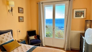 Une chambre à coucher avec un lit, une chaise, une table de chevet, une lampe, un ventilateur, une fenêtre donnant sur un beau paysage et un tableau accroché au mur.